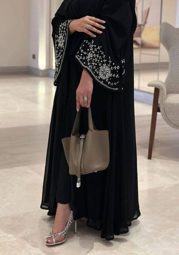 A lower view of the Chiffon-Abaya Schwerelosigkeit, a black abaya with intricate patterns around the hand area, paired with an elegant bag for a refined look.