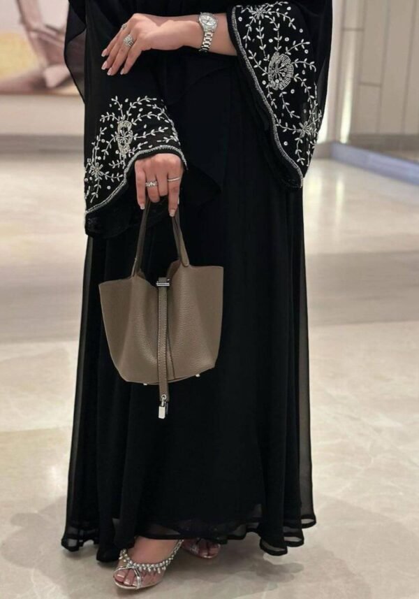 A lady wearing the Chiffon-Abaya Schwerelosigkeit, a black abaya with intricate patterns around the hand area, accessorized with an elegant bag.
