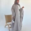 A lady wearing the Lexus Ensemble Vollkommenheit abaya with a light brown exterior and beige interior, walking gracefully next to a wooden bench, accessorized with matching shoes and a silver bag.