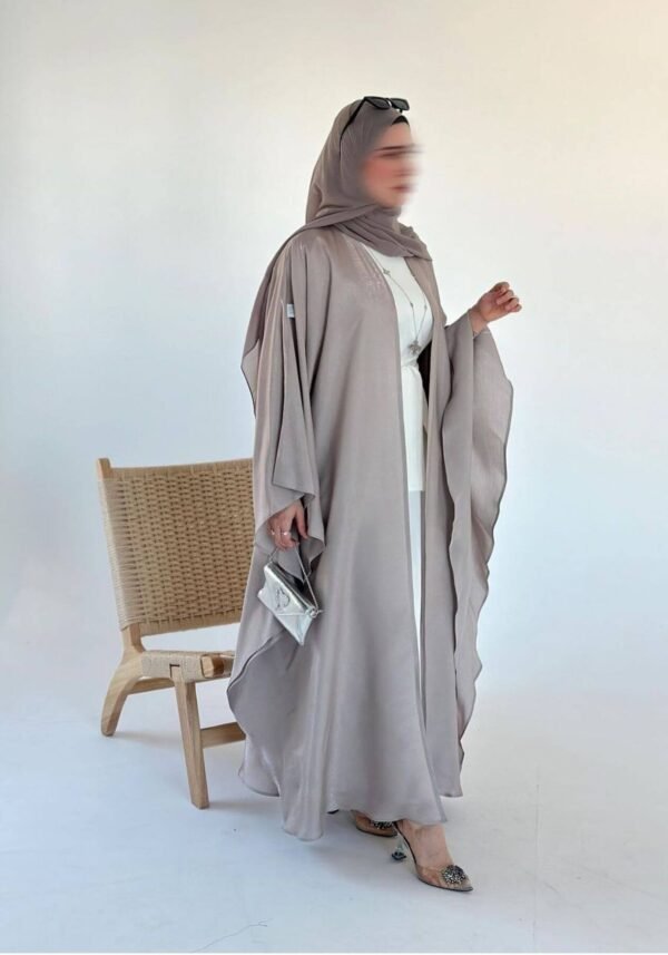 A lady wearing the Lexus Ensemble Vollkommenheit abaya with a light brown exterior and beige interior, walking gracefully next to a wooden bench, accessorized with matching shoes and a silver bag.