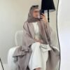 A lady sitting in a salon, wearing the Lexus Ensemble Vollkommenheit abaya with a light brown exterior and beige interior, taking a selfie while accessorized with matching shoes and a silver bag.
