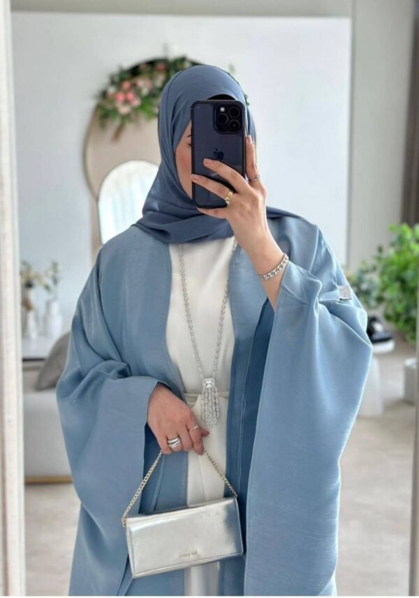 A lady taking a selfie while wearing the Luxus Lexus-Ensemble Royal abaya, featuring a light beige and white interior with a light blue exterior, accessorized with jewelry and a silver bag, having selfie.