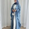 A lady wearing the Luxus Lexus-Ensemble Royal abaya with a light beige and white interior and a light blue exterior, accessorized with jewelry and a silver bag, her face slightly blurred for a graceful, subtle touch.