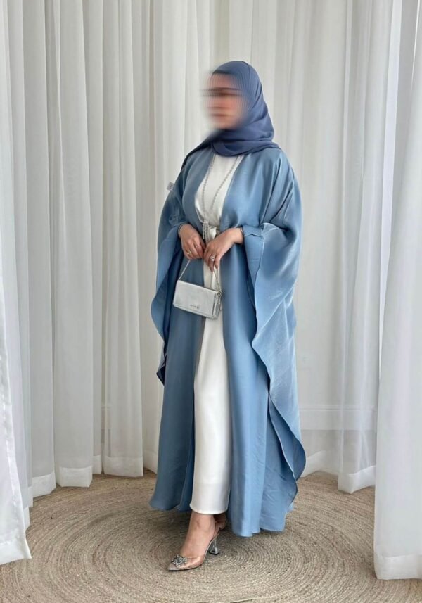 A lady wearing the Luxus Lexus-Ensemble Royal abaya with a light beige and white interior and a light blue exterior, accessorized with jewelry and a silver bag, her face slightly blurred for a graceful, subtle touch.