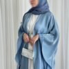 A lady wearing the Luxus Lexus-Ensemble Royal abaya, featuring a light beige and white interior with a light blue exterior, accessorized with jewelry and a silver bag, exuding modern sophistication.