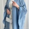 A lady wearing the Luxus Lexus-Ensemble Royal abaya, featuring a light beige and white interior with a light blue exterior, accessorized with jewelry and a silver bag, exuding modern sophistication.
