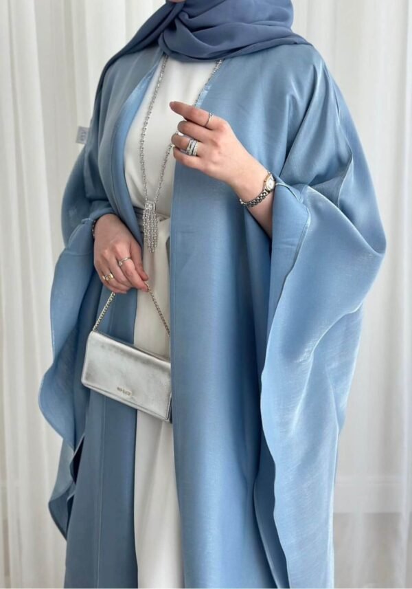 A lady wearing the Luxus Lexus-Ensemble Royal abaya, featuring a light beige and white interior with a light blue exterior, accessorized with jewelry and a silver bag, exuding modern sophistication.