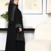 A young lady wearing the Premium Krepp-Abaya Tradition, a black abaya with elegant border patterns, seated in a luxury living room, complemented by a matching bag and shoes ensemble.