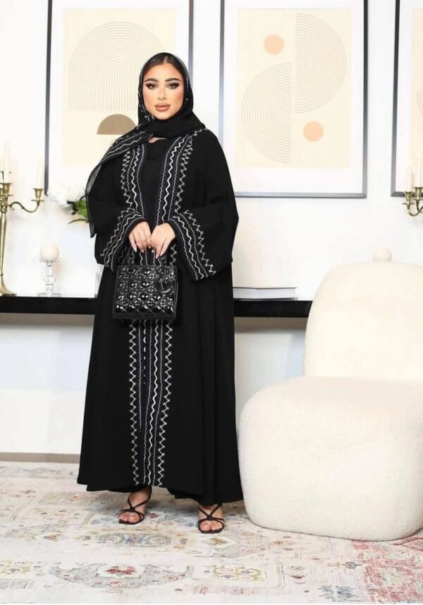 A young lady wearing the Premium Krepp-Abaya Tradition, a black abaya with elegant border patterns, seated in a luxury living room, complemented by a matching bag and shoes ensemble.
