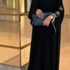 A lady wearing the Royal Chiffon-Abaya Leichtigkeit, a black abaya adorned with elegant jewelry, standing gracefully in a mirror-lined hallway while holding a Yves Saint Laurent bag.