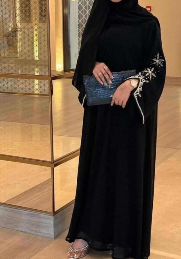 A lady wearing the Royal Chiffon-Abaya Leichtigkeit, a black abaya adorned with elegant jewelry, standing gracefully in a mirror-lined hallway while holding a Yves Saint Laurent bag.