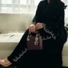 A lady wearing the Royal Krepp-Abaya "Première," featuring silver patterns, seated gracefully on a beige sofa in a living room, holding a stylish bag.
