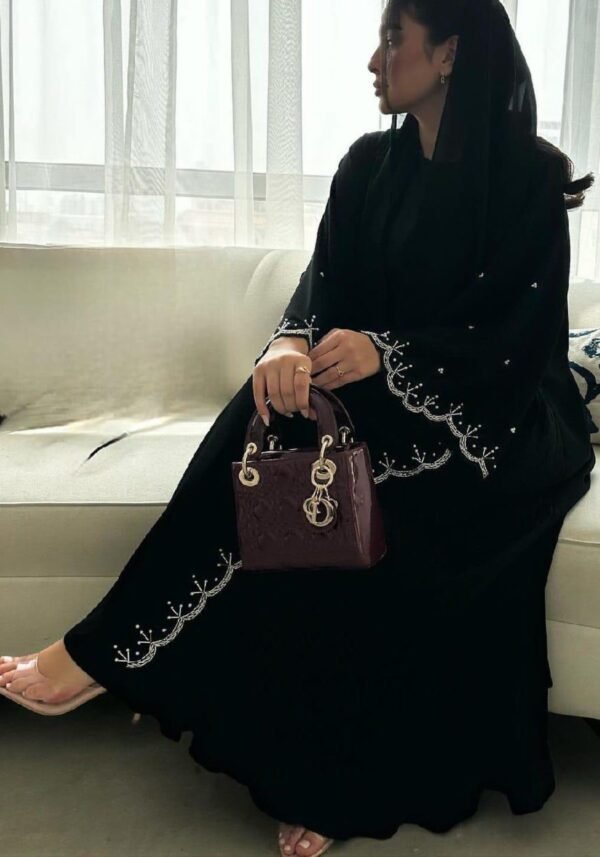 A lady wearing the Royal Krepp-Abaya "Première," featuring silver patterns, seated gracefully on a beige sofa in a living room, holding a stylish bag.
