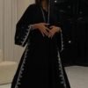 A lady wearing a luxurious abaya with silver patterns, in a living room, holding a stylish bag.