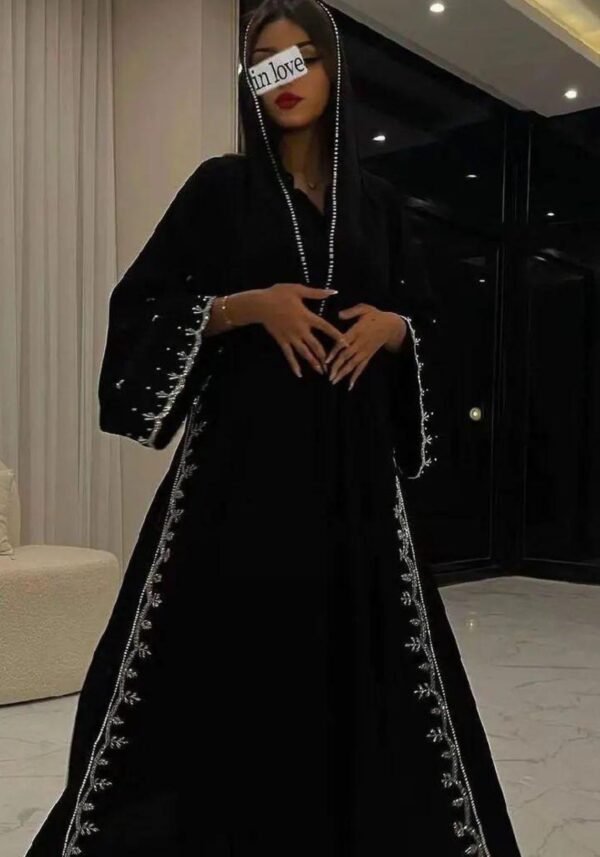 A lady wearing a luxurious abaya with silver patterns, in a living room, holding a stylish bag.