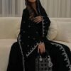 A lady wearing a luxurious abaya with silver patterns, seated gracefully on a beige sofa in a living room, holding a stylish bag.