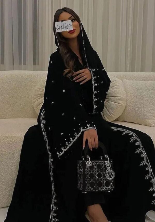 A lady wearing a luxurious abaya with silver patterns, seated gracefully on a beige sofa in a living room, holding a stylish bag.