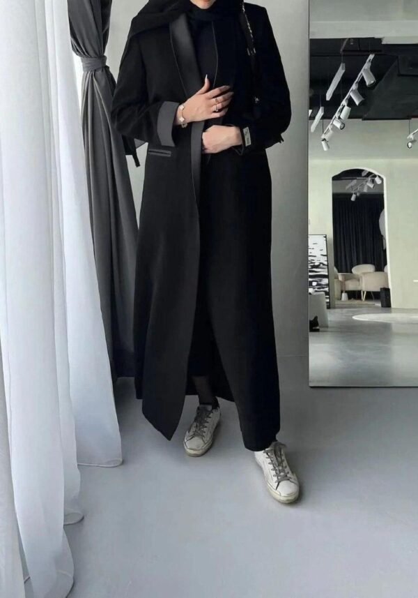 A lady wearing the Klassische Krepp-Abaya Eleganz, standing gracefully next to a modern, simple mirror, highlighting the abaya's timeless elegance.