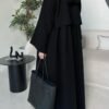 A back view of a lady wearing the Krepp-Abaya Essenz, an all-black abaya paired with a black bag and sporty sneakers, standing in a living room setting.