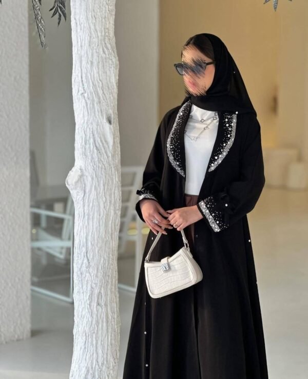 Die Royal Krepp-Abaya mit funkelnden Details, kombiniert mit einer weißen Handtasche, präsentiert in einem modernen und minimalistischen Ambiente.