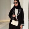 Eine Frau trägt die Royal Krepp-Abaya mit funkelnden silbernen Verzierungen, kombiniert mit einer weißen Handtasche für einen eleganten Look.