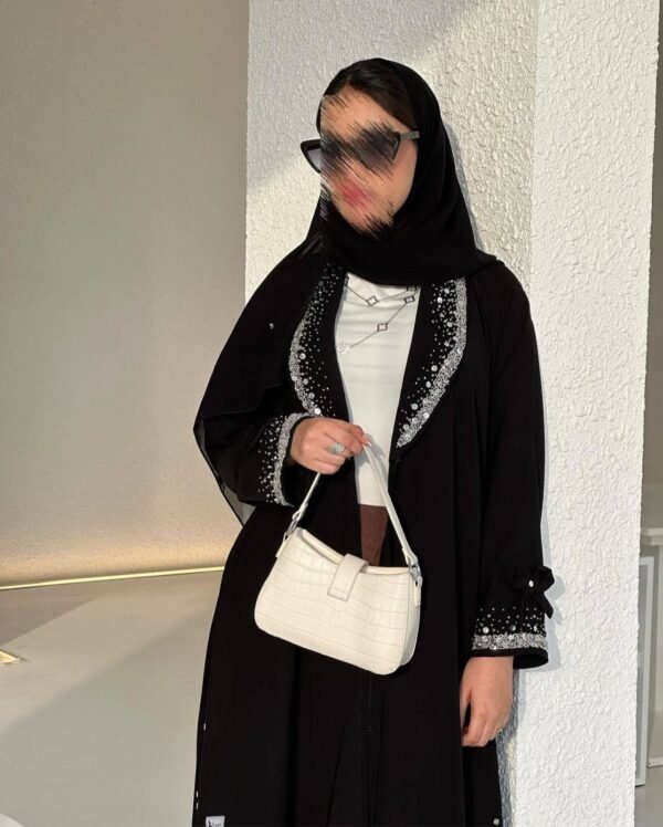 Eine Frau trägt die Royal Krepp-Abaya mit funkelnden silbernen Verzierungen, kombiniert mit einer weißen Handtasche für einen eleganten Look.