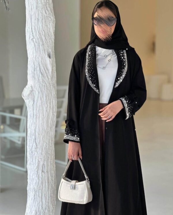 Eine stilvolle Frau in der Royal Krepp-Abaya, mit silbernen Verzierungen und einer weißen Tasche, zeigt die Eleganz dieses klassischen Designs.